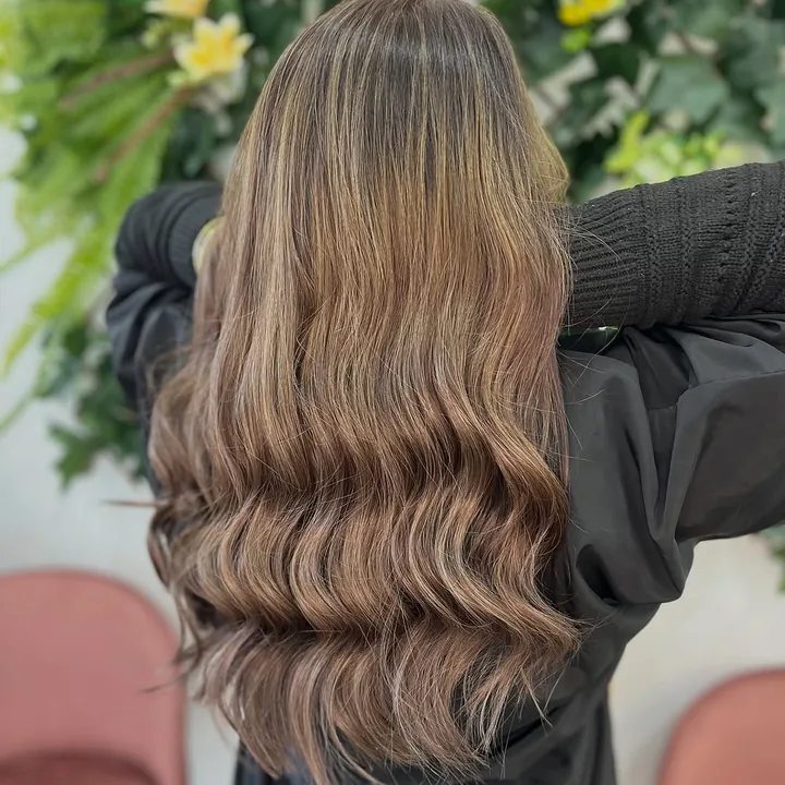Melena bronde en ondas, de espaldas, con la pared de flores y los sillones rosa del sal&oacute;n